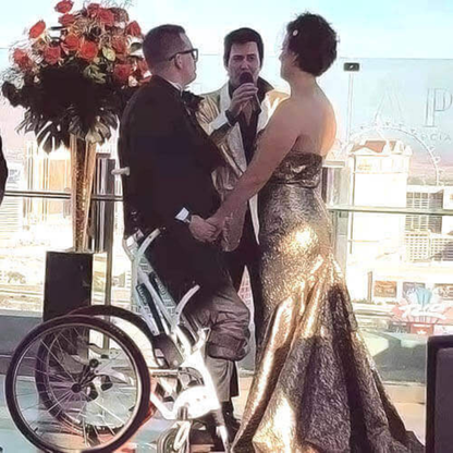 During a wedding ceremony, a couple holds hands while the groom sits in a Foldawheel Lloyd LEO Ultralite Standing Manual Wheelchair. The bride wears a shimmering gown, an Elvis-style officiant speaks, and red flowers decorate the background.