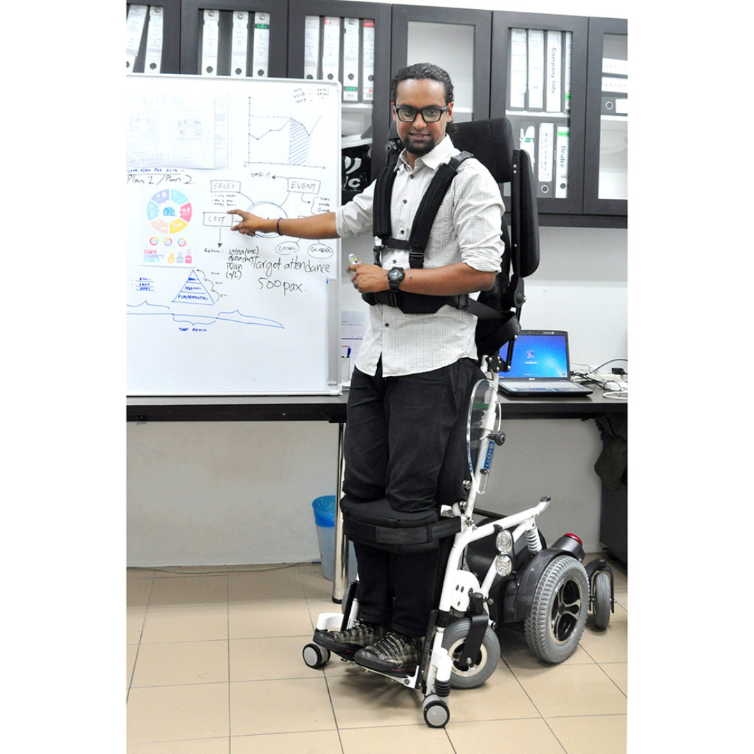 A man using the Foldawheel Lloyd Draco Advanced Standing Power Wheelchair gestures to diagrams and notes on a whiteboard in an office, with shelves of folders and a laptop visible in the background.