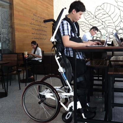 A man uses a Foldawheel Lloyd Pegasus Semi-Powered Standing Wheelchair with a powered sit-to-stand feature at a high café table, typing on his laptop. Around him, others work and drink, with plants and mural art in the background.