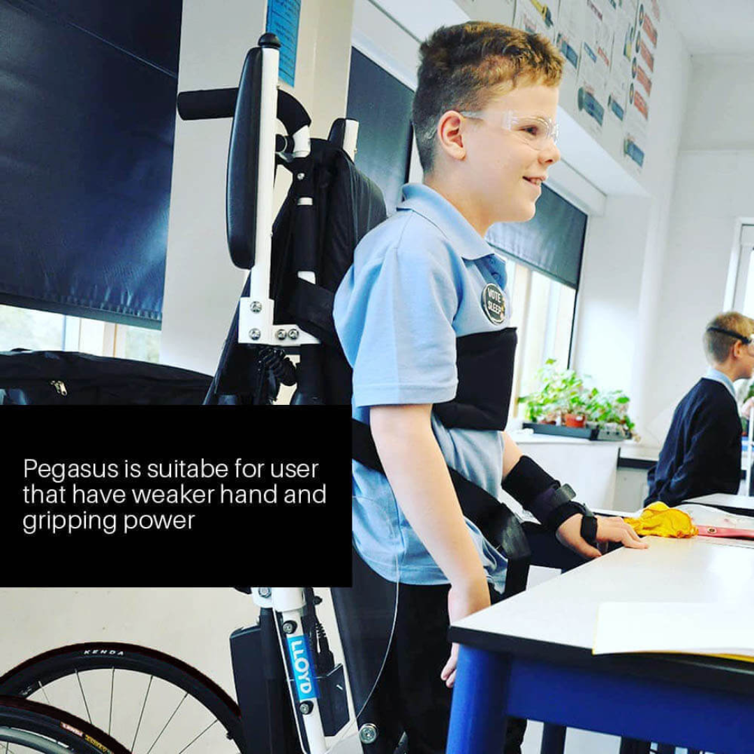 A smiling boy with glasses uses the Foldawheel Lloyd Pegasus Semi-Powered Standing Wheelchair at a classroom desk. Text reads: Pegasus suits users with weaker hand and gripping strength. Another student is visible in the background.