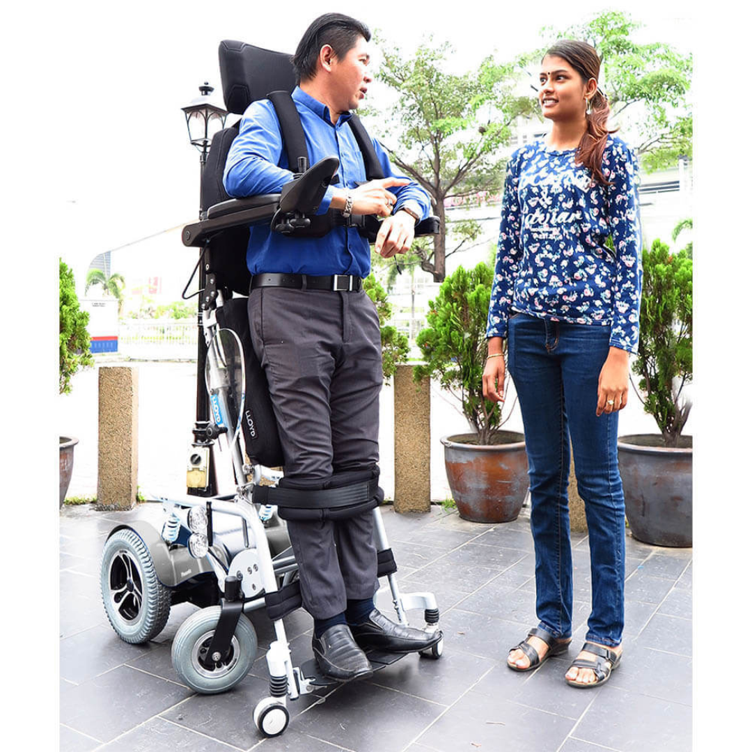 A man in a Foldawheel Lloyd Phoenix 2 Standing Power Wheelchair, which lets him stand, chats with a woman outdoors among plants and trees.