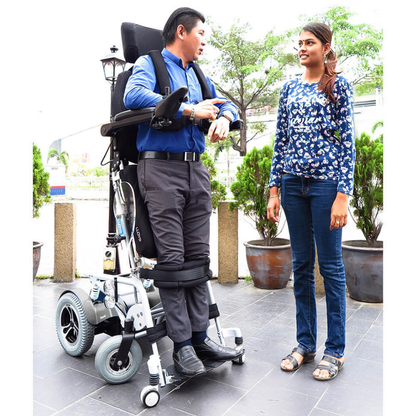 A man in a Foldawheel Lloyd Phoenix 2 Standing Power Wheelchair, which lets him stand, chats with a woman outdoors among plants and trees.