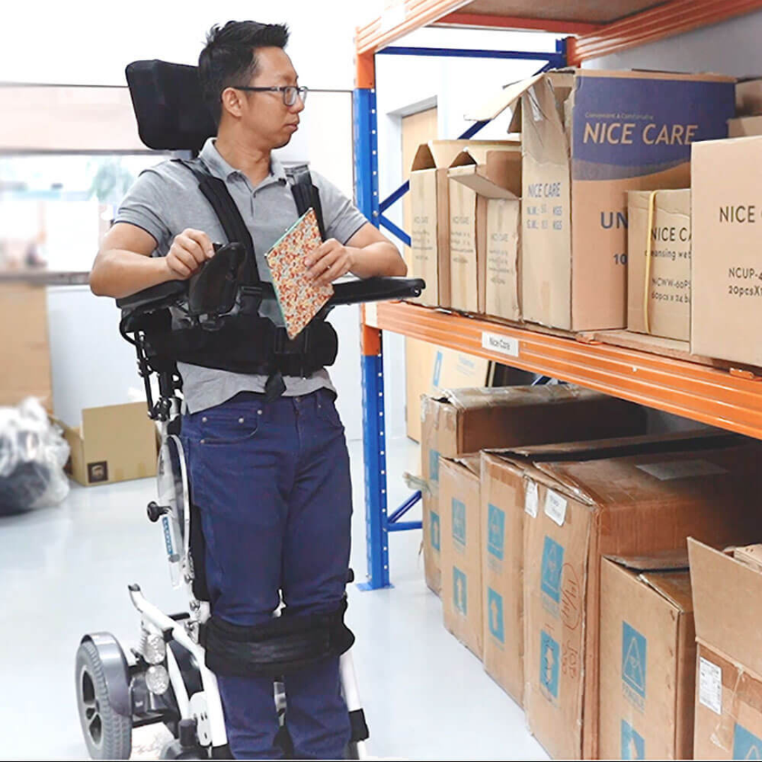 A man in a Foldawheel Lloyd Phoenix 2 Standing Power Wheelchair selects a box from a warehouse shelf while holding a notepad, surrounded by shelves and boxes in the storage area.