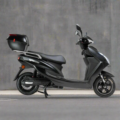 The GIO Phoenix Premium Electric Scooter, an eco-friendly black e-scooter with a rear cargo box, is parked on gray asphalt beside a plain concrete wall.