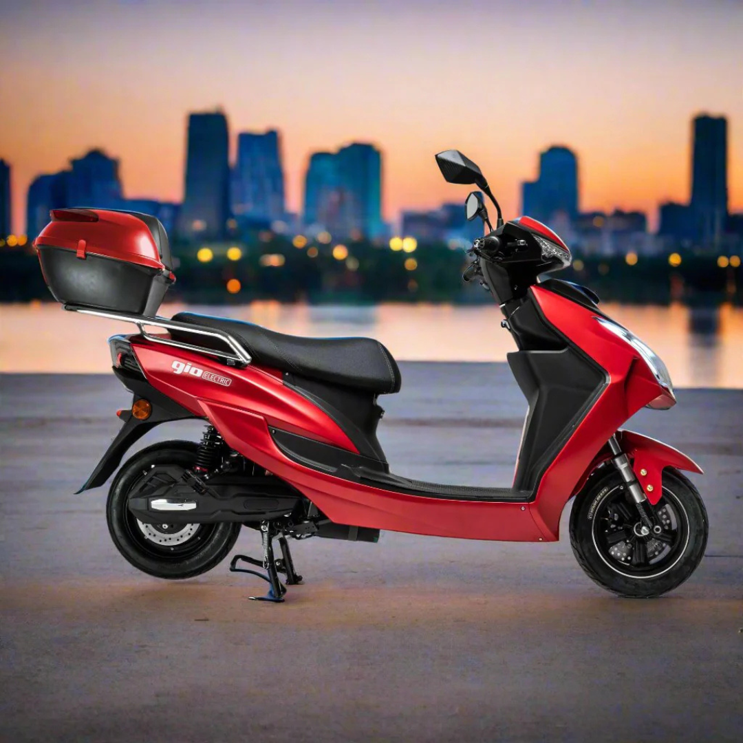 A red GIO Phoenix Premium Electric Scooter with a rear storage box is parked on pavement against a city skyline at sunset. No license needed—ride and enjoy the view.