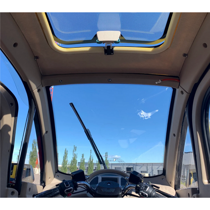 Interior of the GIO Golf Fully Enclosed Mobility Scooter features a clear windshield and sunroof showcasing a blue sky. The dashboard includes navigation and controls. Outside, trees and buildings are visible under the bright sky, providing an enjoyable ride with its heater and cooling fan.