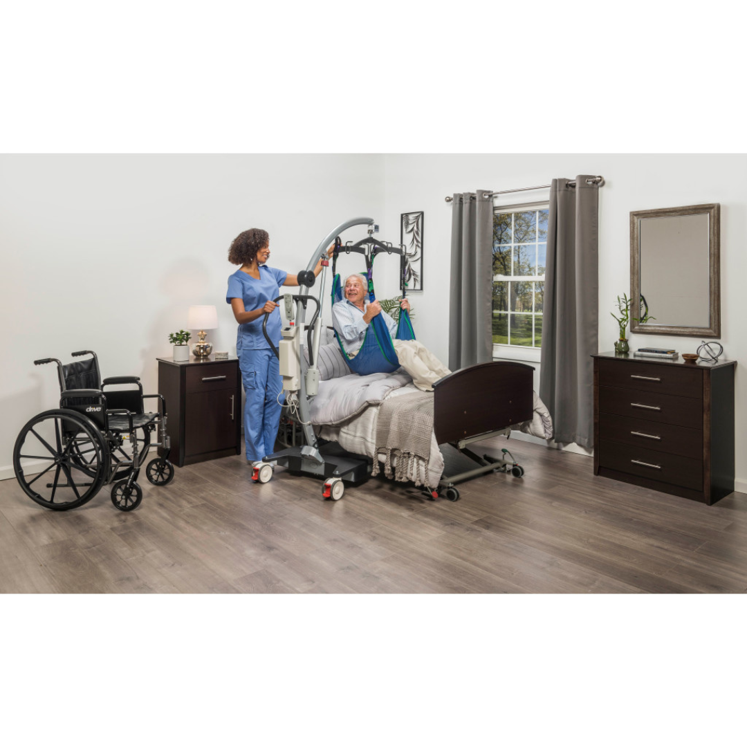 A nurse assists a smiling elderly woman in a hospital bed using the Drive Gravis Battery-Powered Patient Floor Lift. A manual base and wheelchair are nearby in the bright, modern room with wood floors and a curtained window.