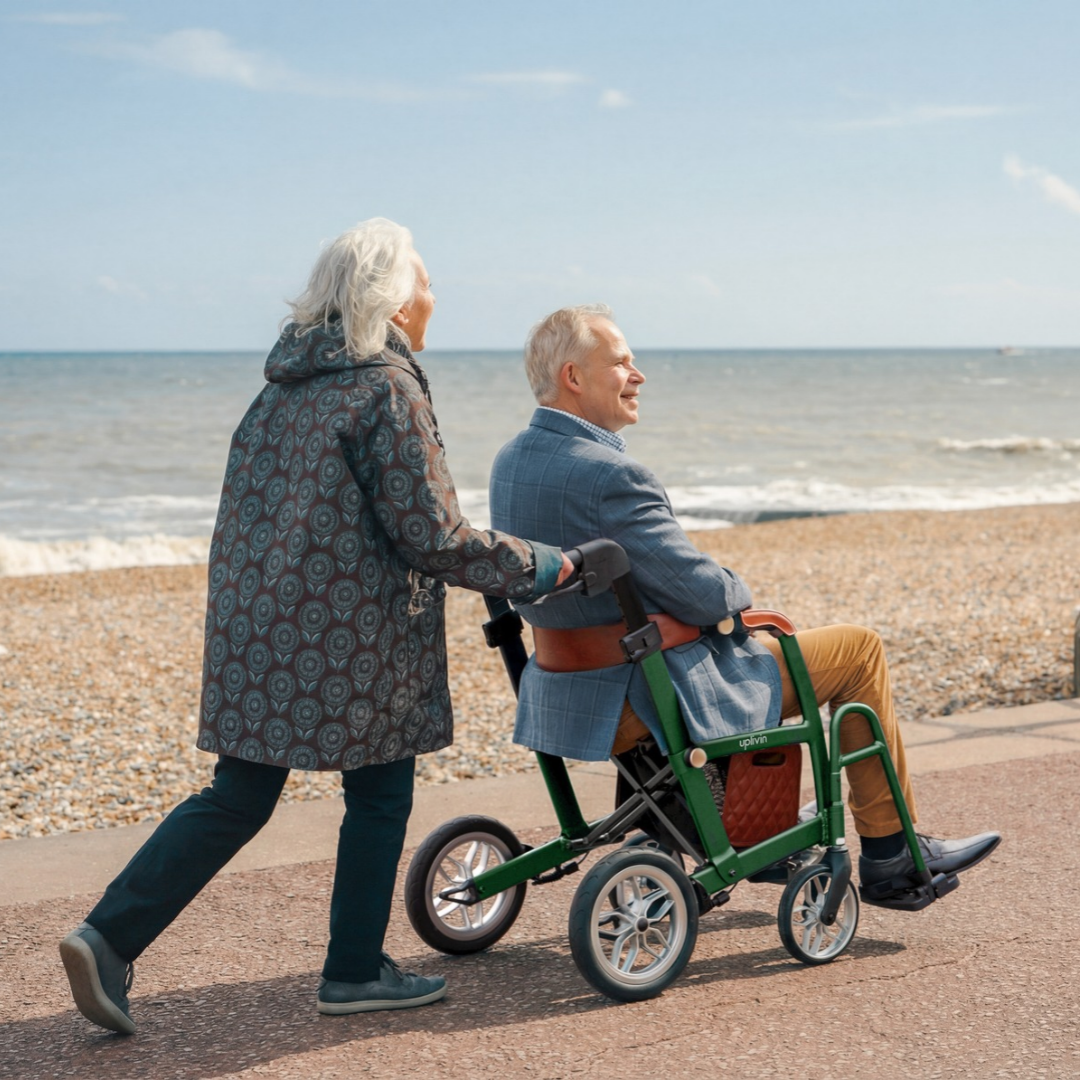 A woman pushes a man sitting in an uplivin Gavo 2-in-1 Hybrid Rollator & Wheelchair along a pebble beach path.
