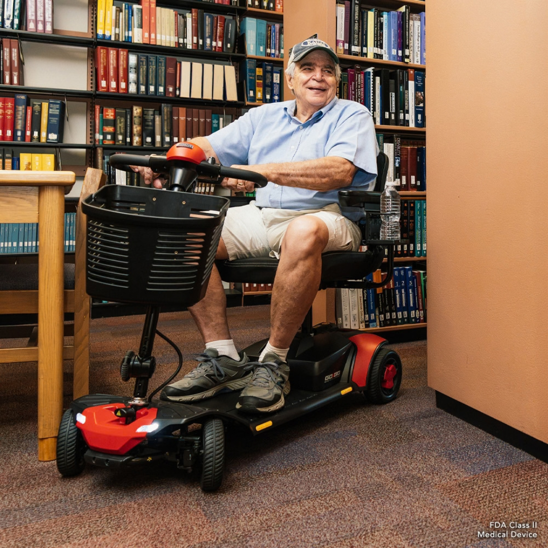 An older man with a baseball cap sits on a Pride Go Go® Elite Traveller Gen 3 Scooter in a library.