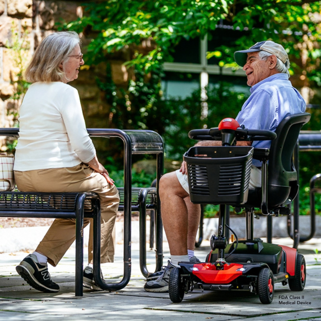 An older man sitting on a red Pride Go Go® Elite Traveller Gen 3 Scooter talks to a woman seated at a patio table outdoors.