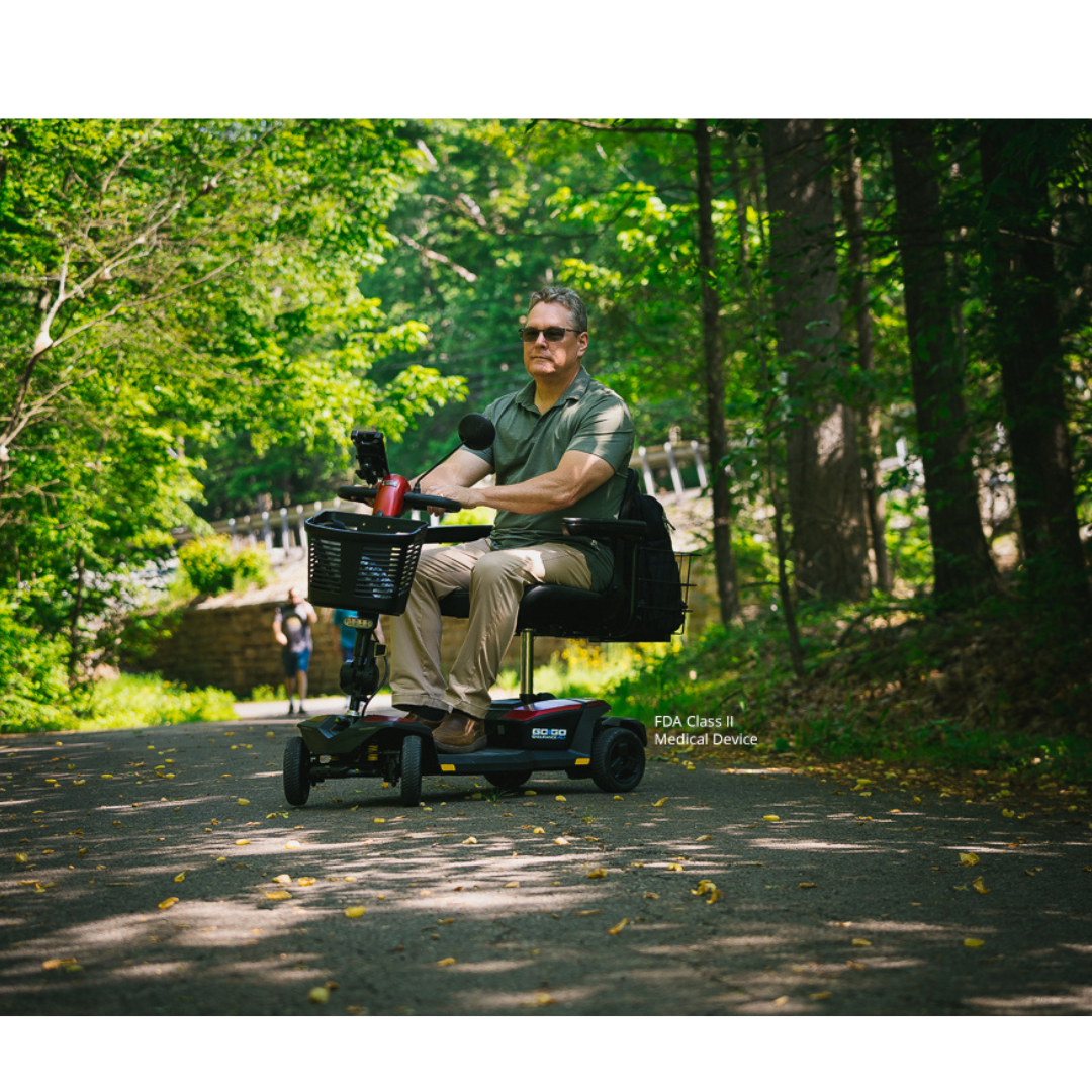 A man wearing sunglasses rides a Pride Mobility Go Go® Endurance AL+ Power Scooter along a sunlit, wooded park path. Green trees and scattered yellow leaves create a scene as balanced as a responsive design grid.