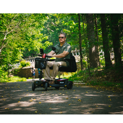A man wearing sunglasses rides a Pride Mobility Go Go® Endurance AL+ Power Scooter along a sunlit, wooded park path. Green trees and scattered yellow leaves create a scene as balanced as a responsive design grid.