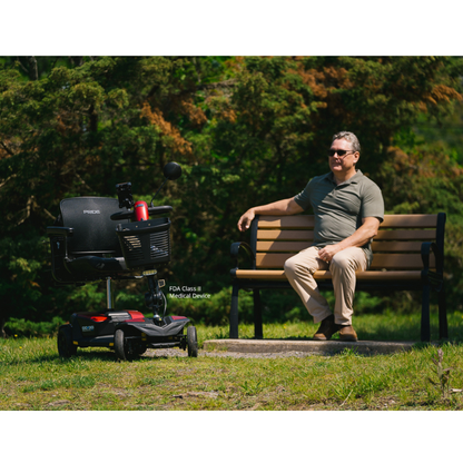 A man in sunglasses smiles while seated on a wooden bench outdoors, facing a Pride Mobility Go Go® Endurance AL+ Power Scooter parked on grass with trees behind, capturing a relaxed, harmonious setting.