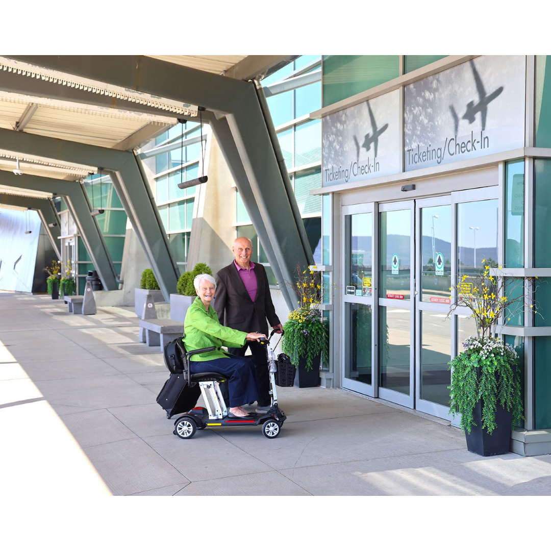 An elderly woman in a green jacket sits on the ultra-portable Golden Technologies Portable Buzzaround GB120 Carry-On Travel Scooter outside an airport terminal, accompanied by a man in a purple sweater. They are near the entrance labeled Ticketing/Check-In with potted plants nearby.