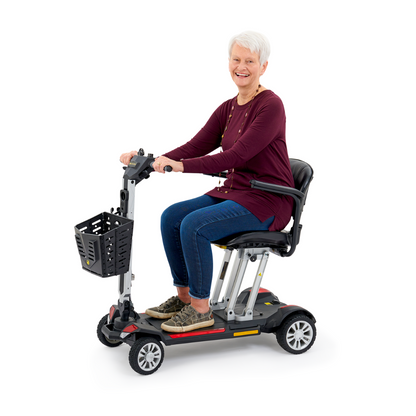 An older adult with short white hair is smiling while seated on a Golden Technologies Portable Buzzaround GB120 Carry-On Travel Scooter. This ultra-portable scooter, powered by a lithium-ion battery, boasts a black basket at the front. The individual is dressed in a dark red top and jeans, perfectly embodying comfort and convenience in motion.
