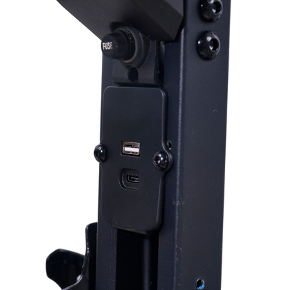 Close-up of a black metal device on the Golden Buzzaround GB220 CarryOn HD Travel Scooter, showing a USB-A port and USB-C port on a rectangular panel, with a fuse button and screws visible above and around the ports.