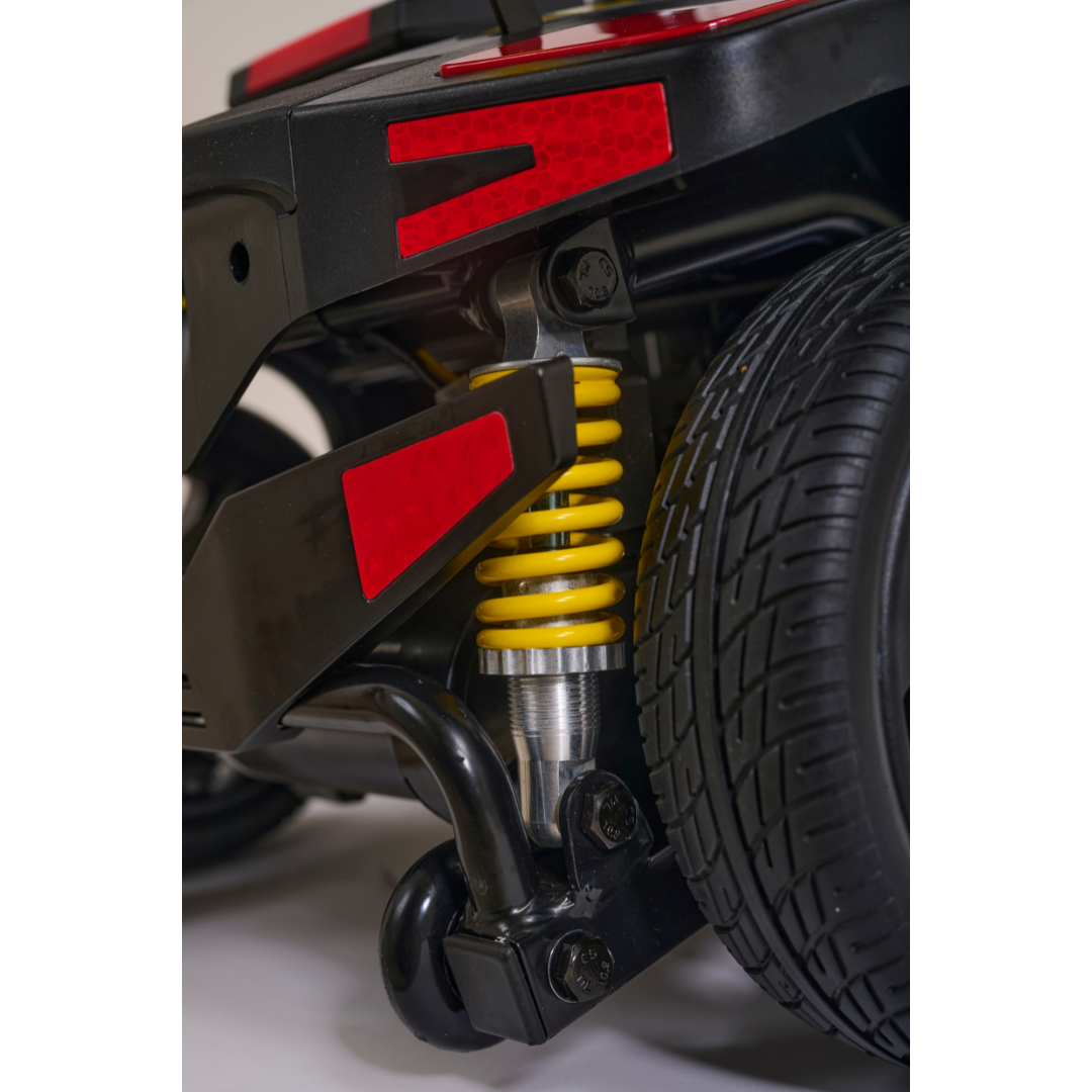 Close-up of the Golden Tech Buzzaround GB152 XL+ 4-Wheel Mobility Scooter’s rear suspension, highlighting its yellow coil spring, black metal parts, red reflectors, and a section of the thick treaded tire.
