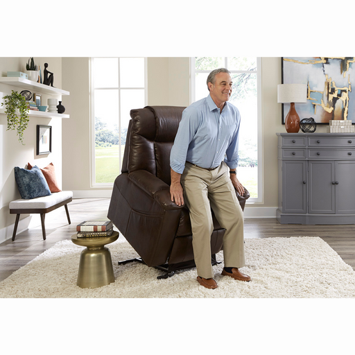 A man in a blue shirt and beige pants uses a brown Golden Tech Rhea PR442 MaxiComfort Recliner with Lift Assist & HeatWave to stand up in a modern living room. The room has a gray sideboard, abstract art, and a small brass table on a white rug, while the recliners USB Charging Port enhances convenience.