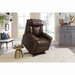 A brown Golden Tech Rhea PR442 MaxiComfort Recliner with Lift Assist & HeatWave sits on a white shag rug in a living room with large windows, a gray sideboard with decor, and shelves of books and plants. Sunlight pours through the windows, casting warmth across the space.