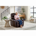 An elderly woman sits in a Golden Cloud+ PR511 Recliner with Lift Assist - Medium Wide, smiling while using a smartphone. The room features modern decor, including a beige rug, a round wooden side table with a mug, plants, and large windows with sheer curtains. A staircase and artwork add charm.