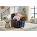 An elderly woman with white hair relaxes in a brown Golden Cloud+ PR511 Recliner with Lift Assist - Medium Wide, adjusting with the AutoDrive hand control for comfort. Dressed in a pink sweater and blue jeans, she is surrounded by modern decor, greenery, a round side table, stairs, and large sunlit windows.