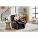 A woman with gray hair sits in a cozy Golden Cloud+ PR511 Recliner with Lift Assist - Medium Wide, holding a remote and drink. Dressed in a pink top and beige pants, shes in a living room featuring a 4-Zone Comfort Zone round side table, houseplants, staircase, and large windows.