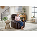An elderly woman with gray hair and glasses relaxes in her Golden Cloud+ PR511 Recliner with Lift Assist - Medium Wide, reading a book. A wooden side table holds a plant and a drink beside her. The bright room is modern, showcasing a staircase and large window.