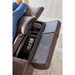 A person sits on a medium wide Golden Cloud+ PR511 Power Lift Recliner, brown leather with a built-in control panel. The open center console shows a remote and smartphone, with adjacent closed compartments. Worn blue jeans, feet hidden. A neutral-toned rug complements the 4-Zone Comfort Zone experience.