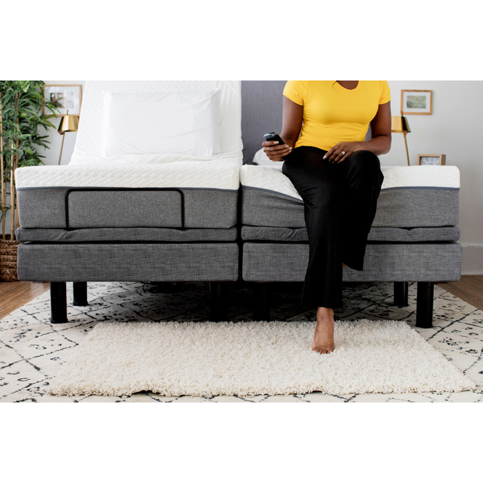 A person in a yellow shirt and black pants sits on the edge of a Golden Passport Hi/Low Full Electric Homecare Bed with Massage, using a remote for video downloads. The room includes a fluffy white rug, framed pictures on the walls, and a potted plant in the corner.