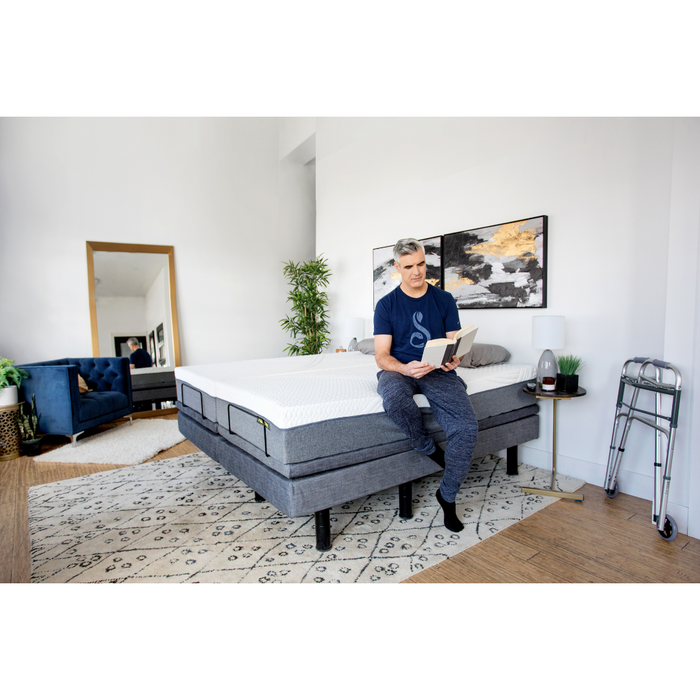 A person relaxes on the Golden Passport Hi/Low Full Electric Homecare Bed with Massage, reading a book amid modern decor. The room includes abstract art, a large mirror, and a dark blue chair. A plant thrives near the walker while online videos softly play in the background, enhancing serenity.