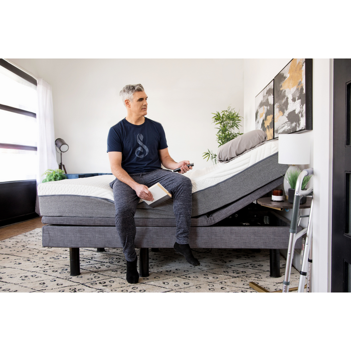 A person relaxes on the edge of a Golden Passport Hi/Low Full Electric Homecare Bed with Massage, holding a book and remote. The bed is slightly elevated, surrounded by a patterned rug, framed art, and a lush plant. Light streams through large windows with sheer curtains as they contemplate downloads or videos next.