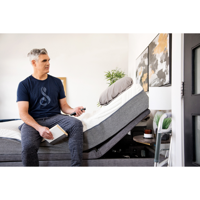 A gray-haired man in a navy shirt and gray pants sits on the edge of a Golden Passport Hi/Low Full Electric Homecare Bed with Massage, remote and notepad in hand, reviewing specs. He gazes towards the headboard. The room features abstract art and a potted plant, exuding thoughtful focus.