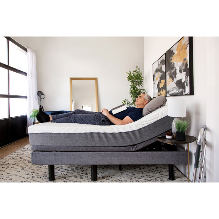 A person relaxes on a Golden Passport Hi/Low Full Electric Homecare Bed with Massage in a modern bedroom, enjoying videos on a tablet. The bed is elevated at the head, flanked by a large mirror, abstract art, a plant, and a lamp. A patterned rug adorns the floor.