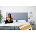 A woman in a yellow shirt and black pants sits cross-legged on the Golden Passport Hi/Low Full Electric Homecare Bed with Massage, adjusting bedding while smiling. The room, ideal for home decor videos, has a gray headboard, gold lamps, framed pictures, and a green plant in the corner.