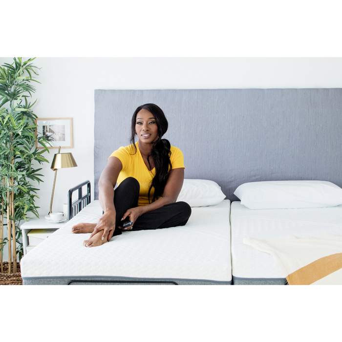 A person relaxes on a Golden Passport Hi/Low Full Electric Homecare Bed with Massage featuring a gray headboard, in a bright, modern bedroom. Dressed in a yellow shirt and black pants, they review video downloads on their laptop amidst lush plants, an elegant lamp, and charming framed art.