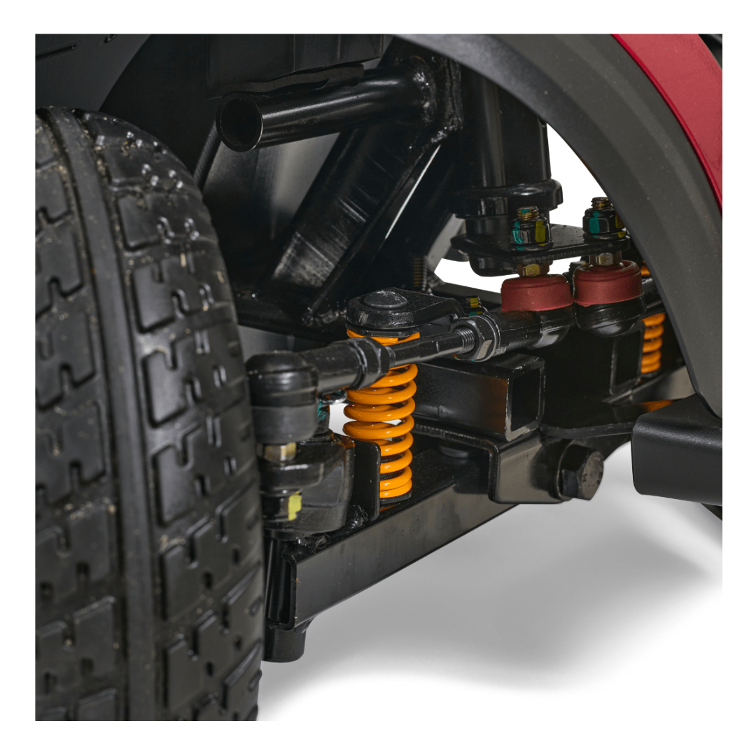 Close-up of an ATV suspension system with orange coil springs and mechanical parts, resembling a Golden Tech GB148 Buzzaround EX Portable Mobility Scooter. A large off-road tire, treaded for rugged terrain, is visible on the left. Components are clean against a white background.