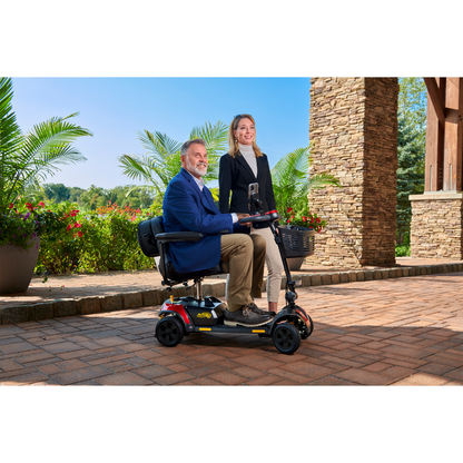 A man in a blue blazer relaxes on his Golden Tech GB124 Buzzaround XL 4-Wheel Portable Mobility Scooter with an extended range battery beside a smiling woman in a black jacket and beige pants. They enjoy the stone patio with large potted plants and a grand stone column.