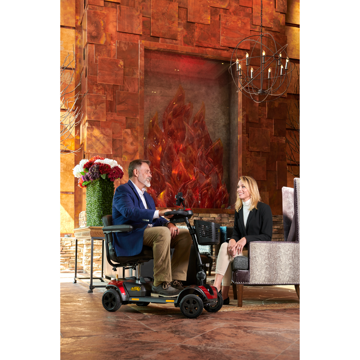 A man on a Golden Tech GB124 Buzzaround XL 4-Wheel Mobility Scooter with an extended range battery talks to a seated woman in a warmly lit modern room featuring a large decorative fire element and a contemporary chandelier.
