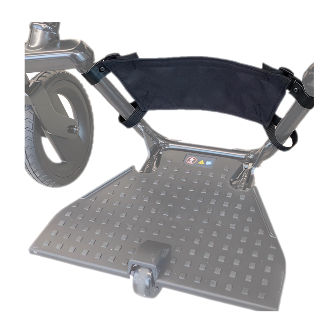 Close-up of the Heel Stopper for WHILL Model F Powerchair, showing its textured, grid-like footrest surface, black fabric calf strap, and a large wheel partially visible on the left side.