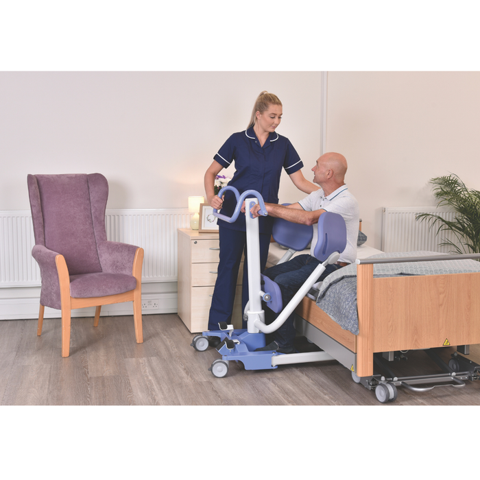 A nurse assists an older man beside a hospital bed using the Joerns Hoyer UP Sit-to-Stand Compact Manual Patient Lift in a well-lit room with a purple armchair, nightstand, and potted plant, supporting his mobility and independence.