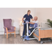 A nurse assists an older man beside a hospital bed using the Joerns Hoyer UP Sit-to-Stand Compact Manual Patient Lift in a well-lit room with a purple armchair, nightstand, and potted plant, supporting his mobility and independence.