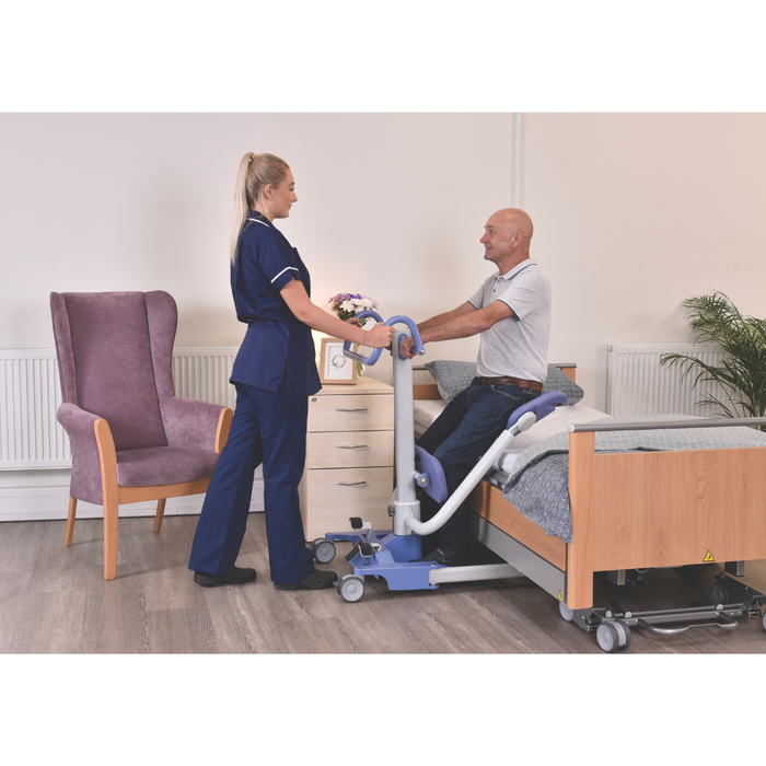 A nurse assists a smiling man with the Joerns Hoyer UP Sit-to-Stand Compact Manual Patient Lift beside a hospital bed in a bright room with a purple chair, cabinet, and plant, promoting mobility and independence.