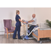 A nurse assists a smiling man with the Joerns Hoyer UP Sit-to-Stand Compact Manual Patient Lift beside a hospital bed in a bright room with a purple chair, cabinet, and plant, promoting mobility and independence.
