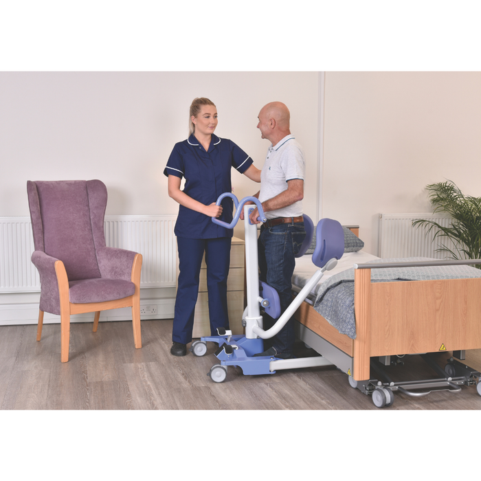 A nurse assists a man using the Joerns Hoyer UP Sit-to-Stand Compact Manual Patient Lift for a weight-bearing transfer beside a hospital bed in a room with a purple armchair and potted plant.