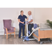 A nurse assists a man using the Joerns Hoyer UP Sit-to-Stand Compact Manual Patient Lift for a weight-bearing transfer beside a hospital bed in a room with a purple armchair and potted plant.