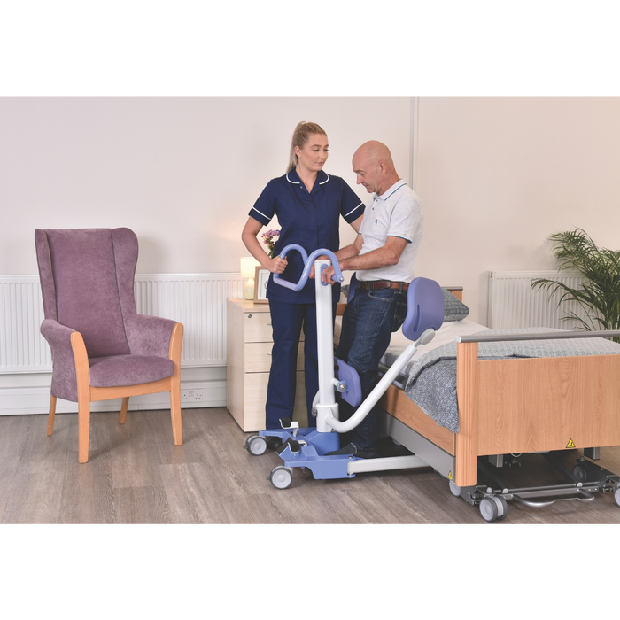 A nurse uses the Joerns Hoyer UP Sit-to-Stand Compact Manual Patient Lift to help an older man stand near a wooden bed in a care home. The room includes a purple armchair, a plant, and light-colored furniture.