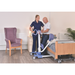 A nurse uses the Joerns Hoyer UP Sit-to-Stand Compact Manual Patient Lift to help an older man stand near a wooden bed in a care home. The room includes a purple armchair, a plant, and light-colored furniture.