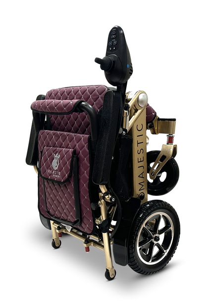 Introducing the MAJESTIC IQ-7000, a compact and foldable electric wheelchair featuring burgundy quilted cushioning and gold metallic accents. This model offers intelligent joystick control on a black handle, along with sleek black wheels and auto-folding capabilities for effortless transportation and storage.