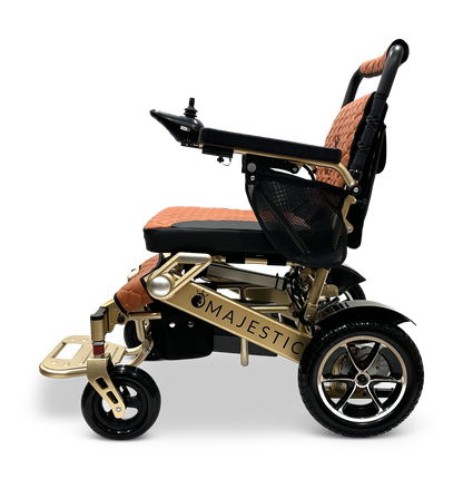 The MAJESTIC IQ-7000 is a gold-colored auto-folding electric wheelchair with brown and black cushioning, featuring a 360-degree joystick on the armrest and small front wheels for easy maneuverability. The lightweight frame displays the name Majestic against a black background.