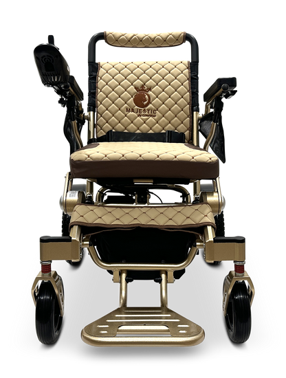 The MAJESTIC IQ-7000 Remote Controlled Electric Wheelchair is a luxurious model in beige and brown, featuring quilted upholstery, armrests, joystick control, and a footrest. The name "Majestic" is elegantly embroidered on the backrest and enhanced with sleek gold-tone accents. This lightweight folding wheelchair design ensures ease during travel.